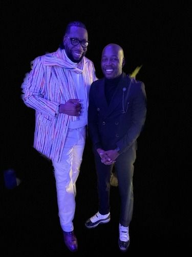 Two people posing together in dark clothes under purple stage lighting, smiling at the camera.