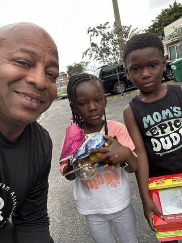 Man and two children smiling outdoors, holding snacks near parked cars