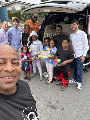 Group posing with gifts beside an open SUV on a residential street, with one person taking a selfie in front.