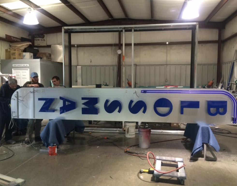 People working on a large illuminated sign indoors. The sign says
