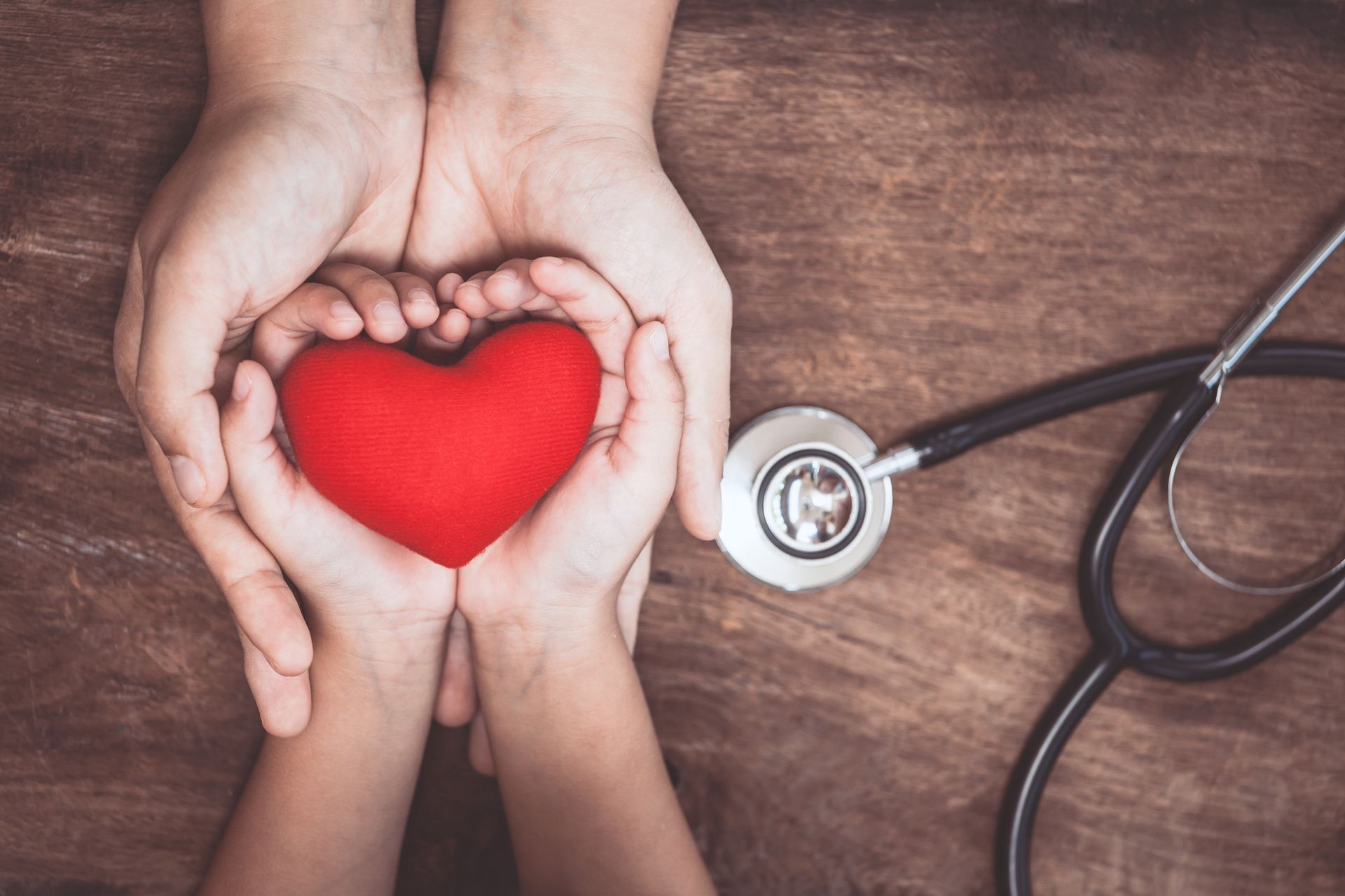 A child's hands is holding a toy heart and below are adult hands. There is a stethoscope next to the hands.