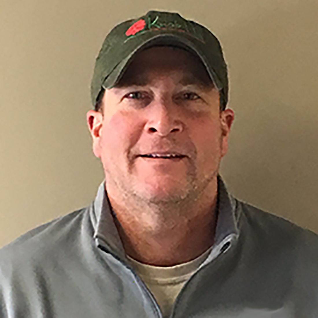 Smiling man in a green cap and gray jacket against a plain beige wall