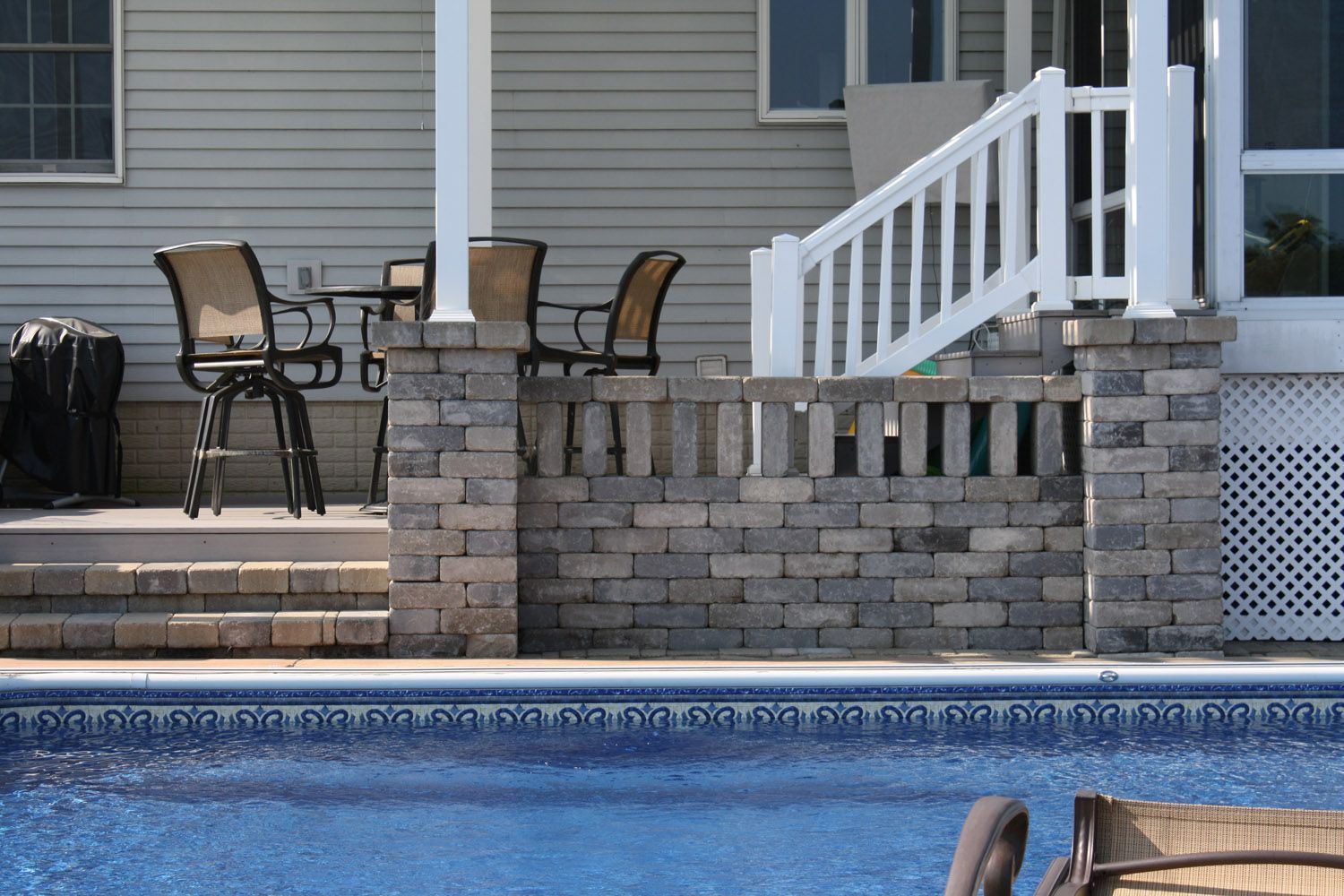Patio chairs and table beside a backyard pool with brick retaining wall and house steps
