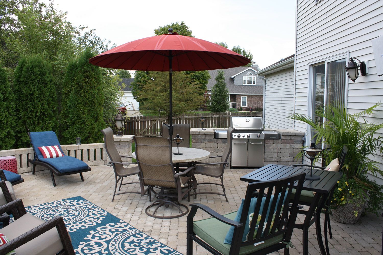 Backyard patio with red umbrella, outdoor dining set, grill, lounge chairs, and potted plants