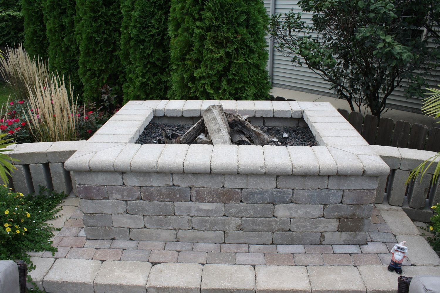 Stone backyard fire pit with square paver top, gravel inside, and landscaped shrubs behind
