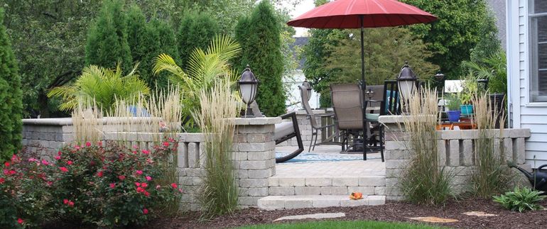 Backyard patio with stone steps, red umbrella, black chairs, and flower beds surrounded by greenery