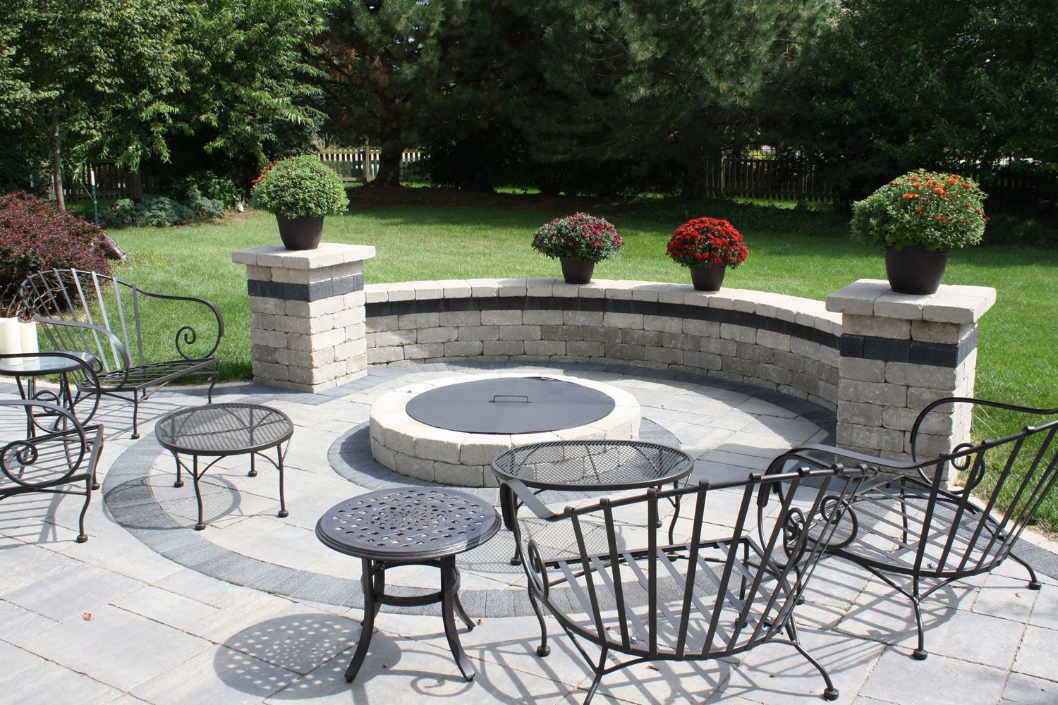 Backyard patio with curved stone fire pit, metal chairs, and potted flowers on benches