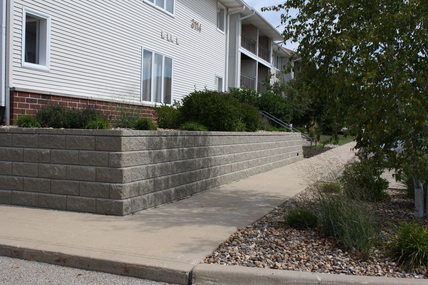 Apartment building entrance with raised concrete retaining wall, sidewalk ramp, and shrubs along the path