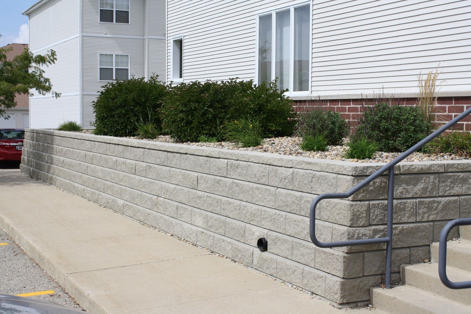 Concrete retaining wall beside a white house, with shrubs and a wheelchair ramp railing on the right.