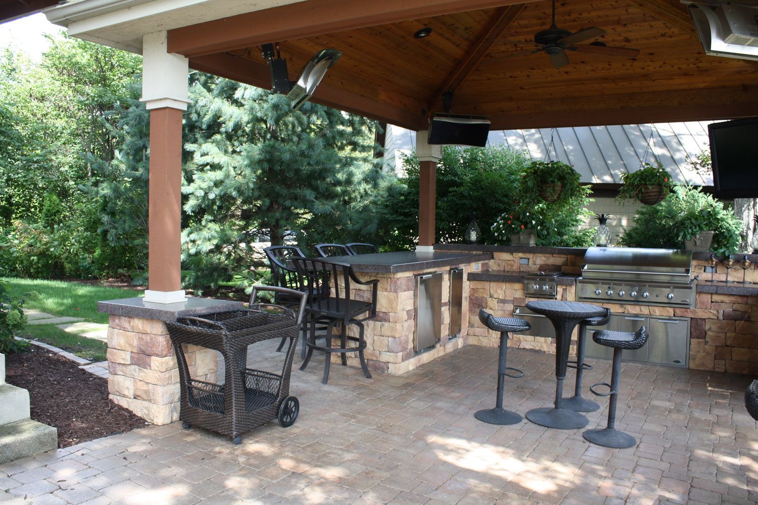 Covered outdoor patio with stone fireplace, bar stools, and a grill under a wooden roof