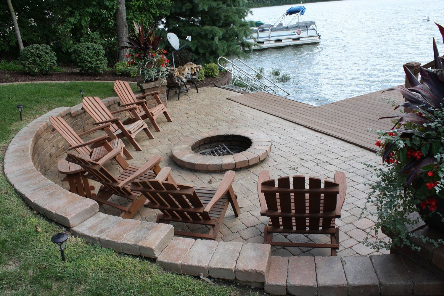 Curved stone fire pit with wooden chairs beside a lakeside dock and boat