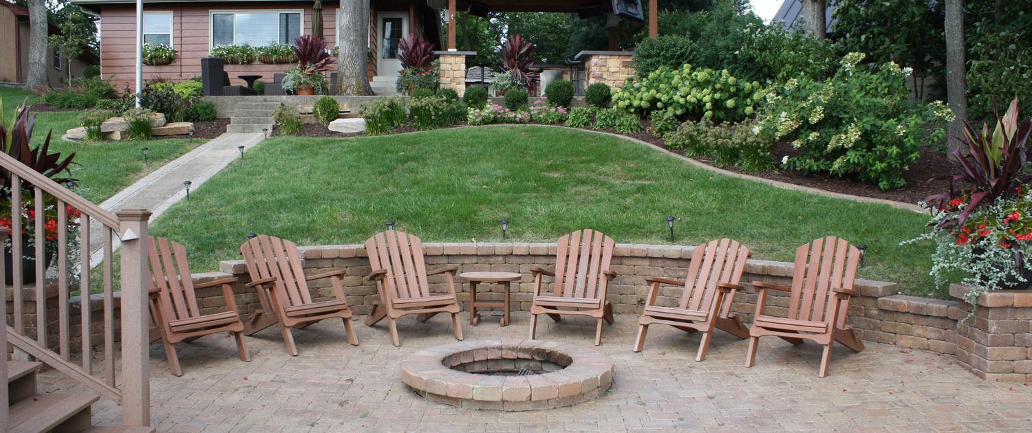 Backyard patio with a circular fire pit surrounded by wooden chairs and a landscaped lawn