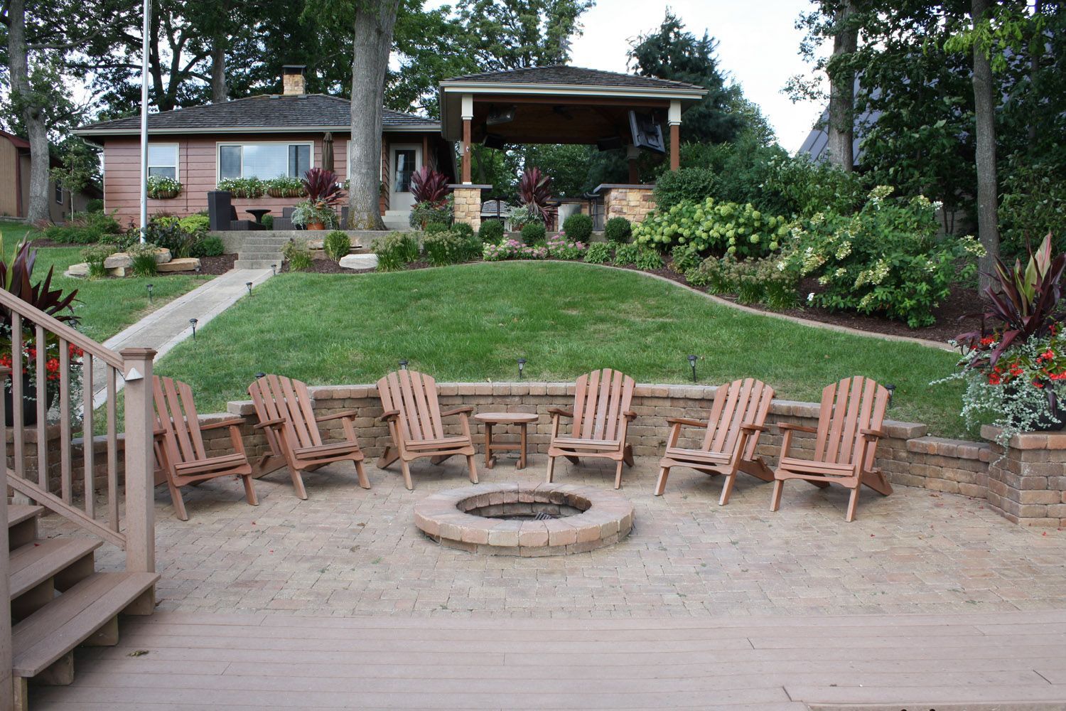 Backyard patio with wooden chairs around a fire pit and a landscaped hill leading to a house