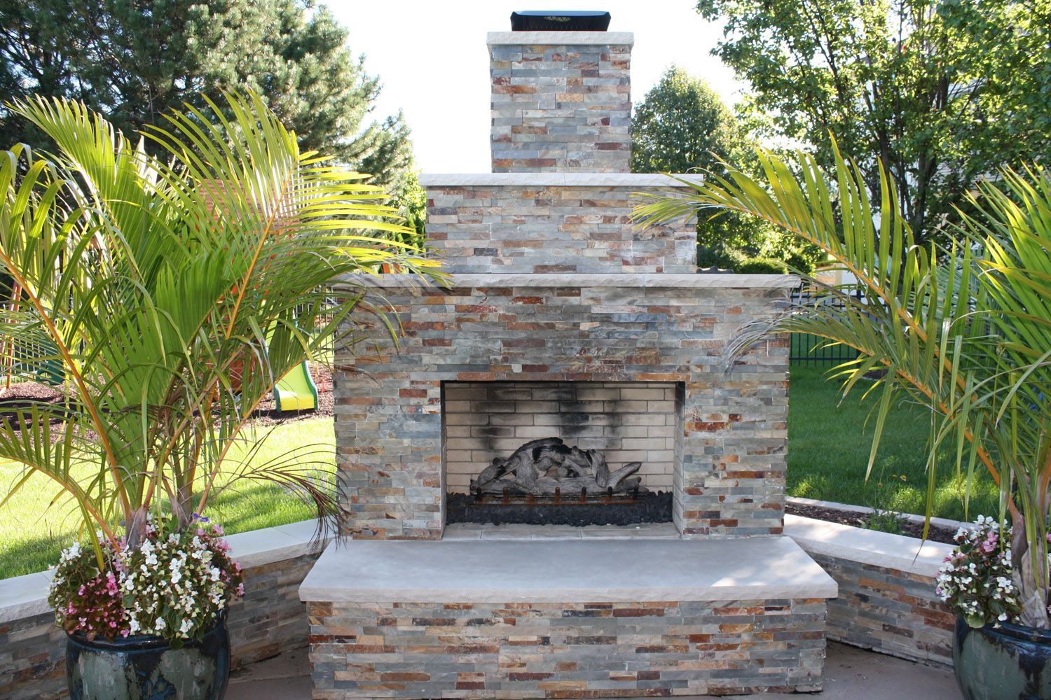 Stone outdoor fireplace with a rectangular firebox, surrounded by palm plants and greenery