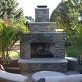 Stone outdoor fireplace with chimney in a landscaped patio, surrounded by greenery and seating