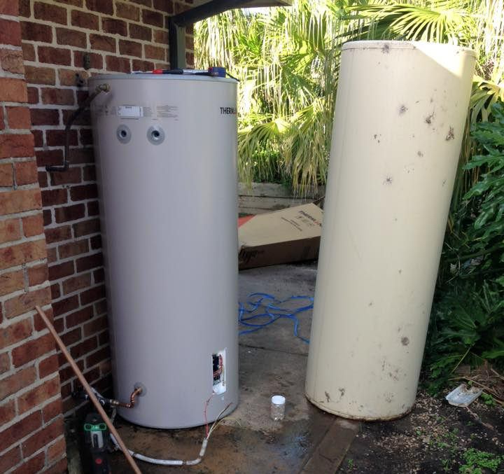 Two large water heaters beside a brick wall, one gray and one beige, in a backyard area. Two large water heaters beside a brick wall, one gray and one beige, in a backyard area.