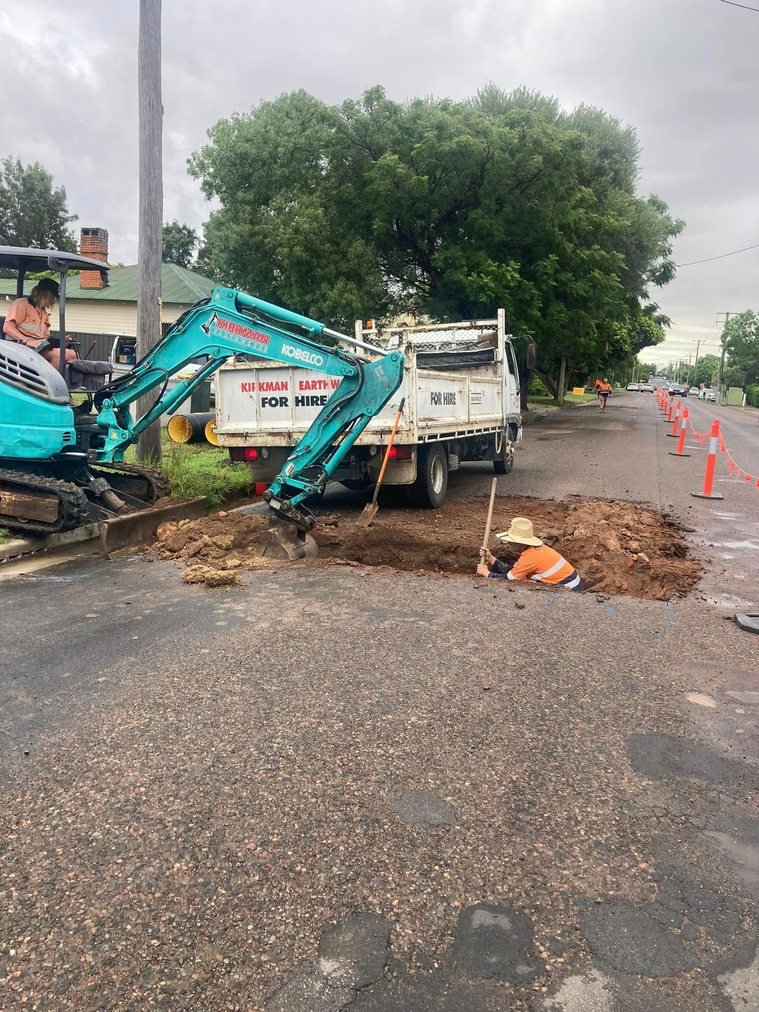 Backhoe loading muddy debris into a white truck beside a wet roadside with orange cones. Backhoe loading muddy debris into a white truck beside a wet roadside with orange cones.