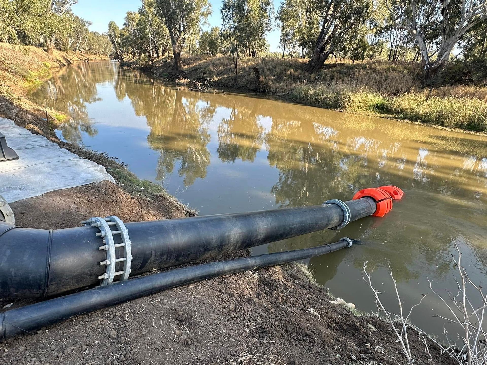 Black pipe with orange valve extending into a muddy canal beside a tree-lined bank Black pipe with orange valve extending into a muddy canal beside a tree-lined bank