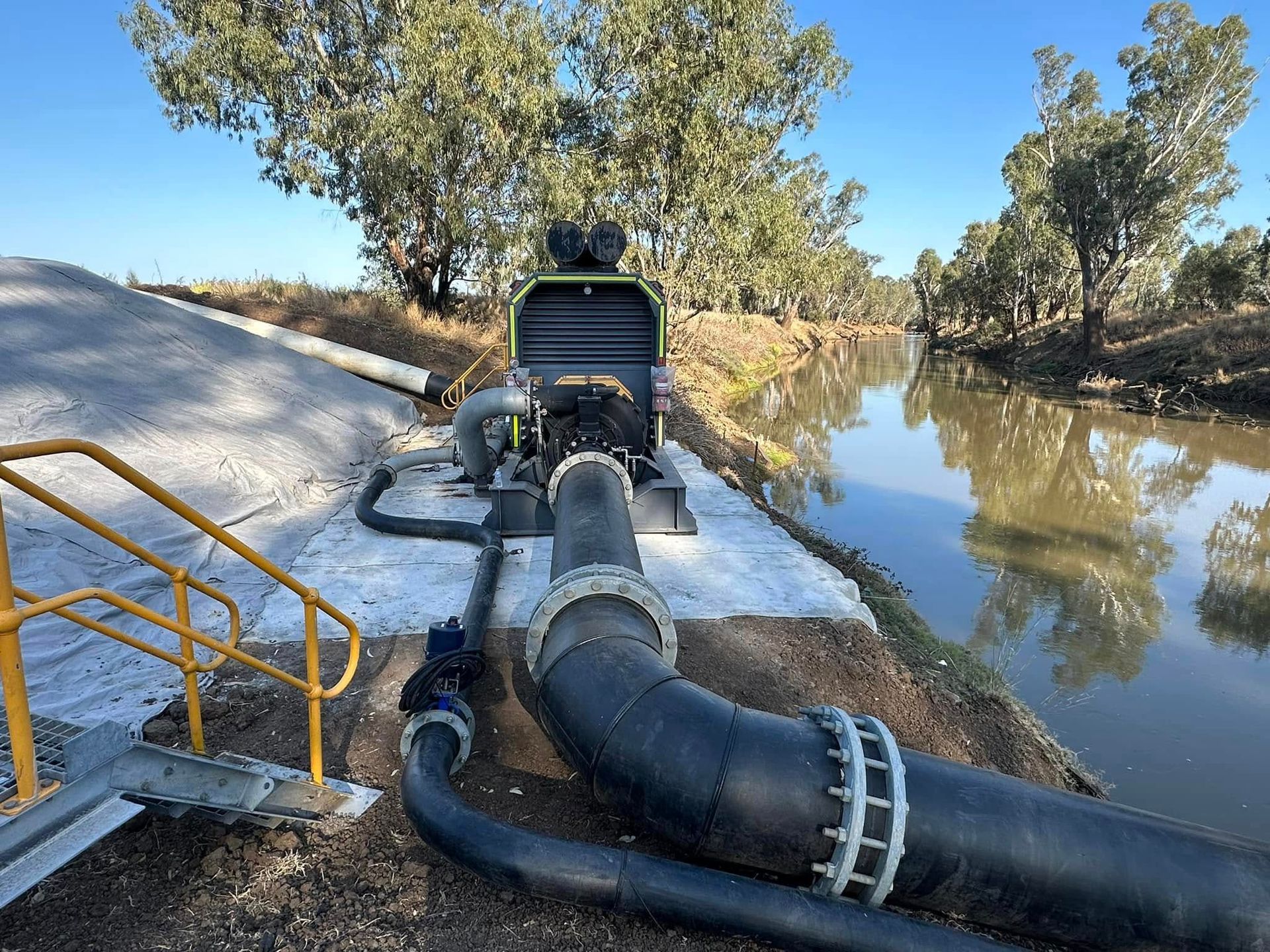 Pump and hoses beside a river, moving water through a dredging setup near trees and a walkway Pump and hoses beside a river, moving water through a dredging setup near trees and a walkway