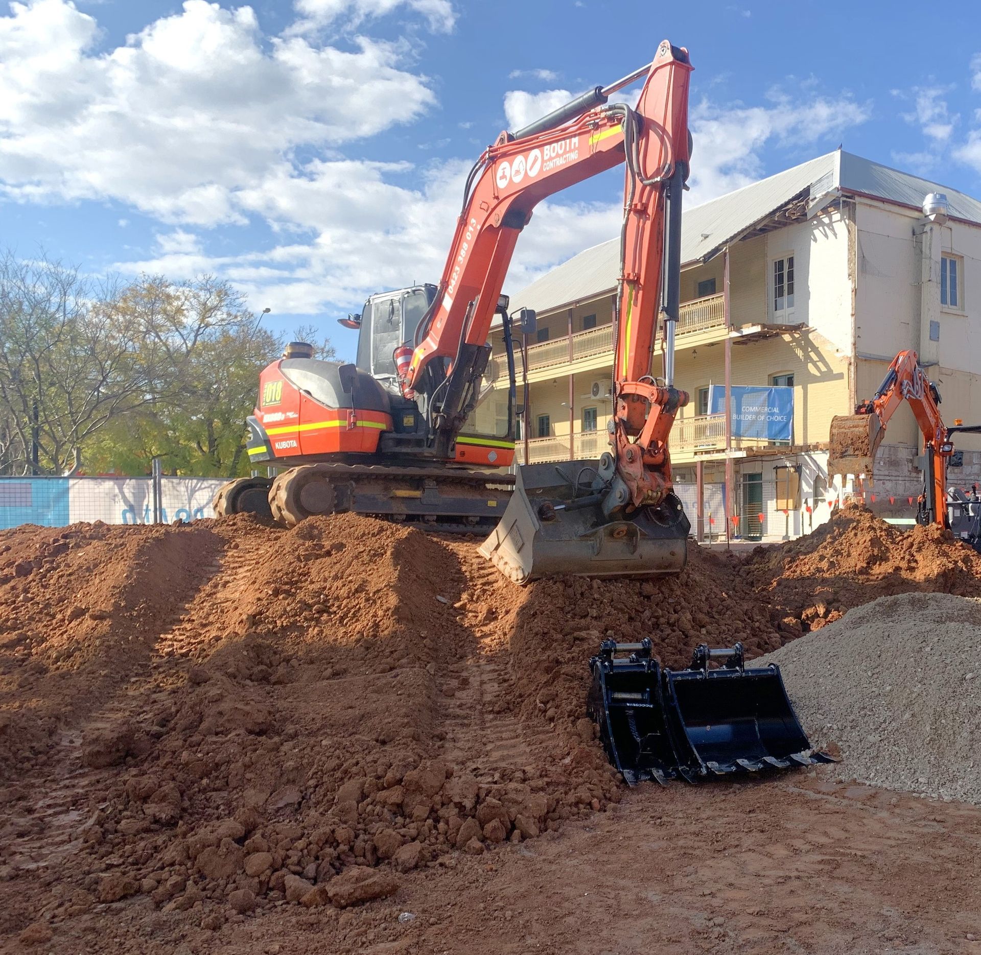 Orange excavator digging dirt at a construction site beside a building under a blue sky Orange excavator digging dirt at a construction site beside a building under a blue sky