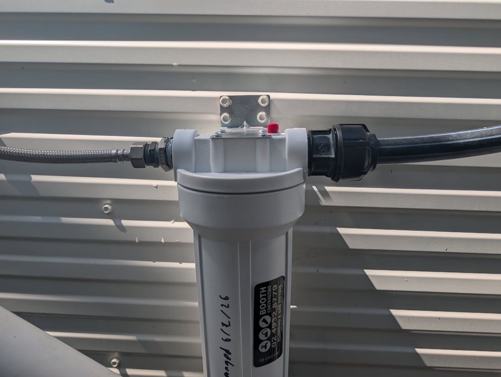 Gray inline water filter installed on a black pipe against a corrugated metal wall. Gray inline water filter installed on a black pipe against a corrugated metal wall.