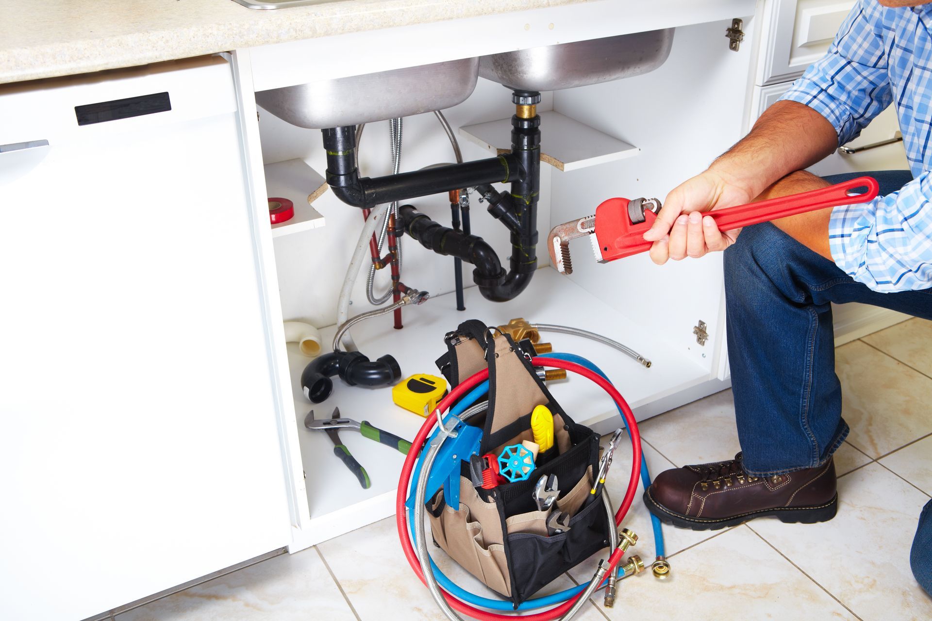 Plumber tightening a sink pipe under a cabinet with tools and open tool bag nearby Plumber tightening a sink pipe under a cabinet with tools and open tool bag nearby