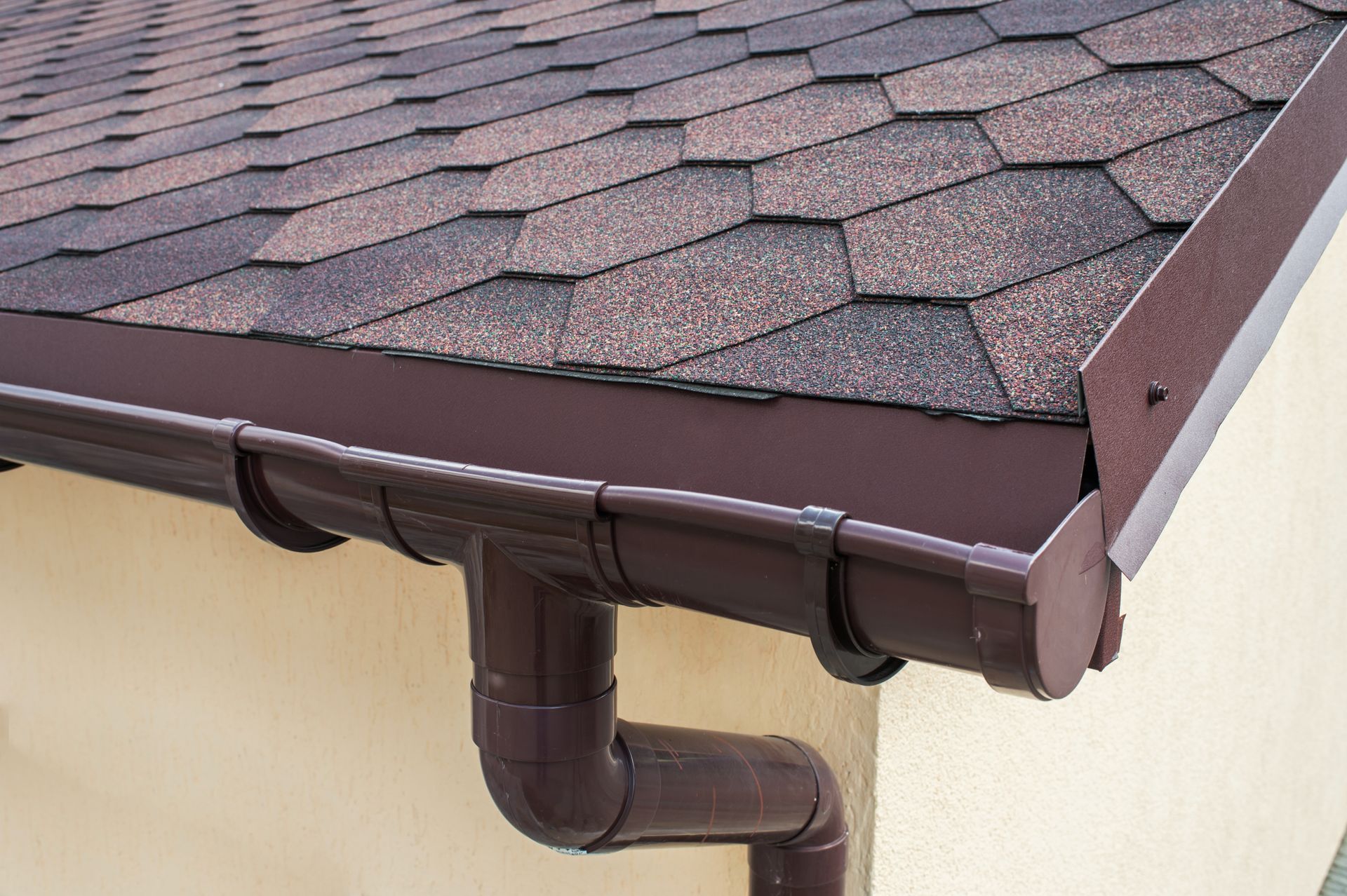 Brown gutter and downspout on a beige house beneath a shingled roof