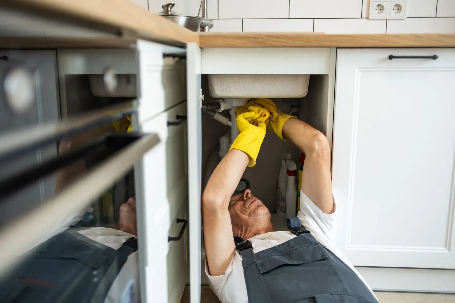 Plumber in yellow gloves repairs a pipe under a kitchen sink while lying on the floor.