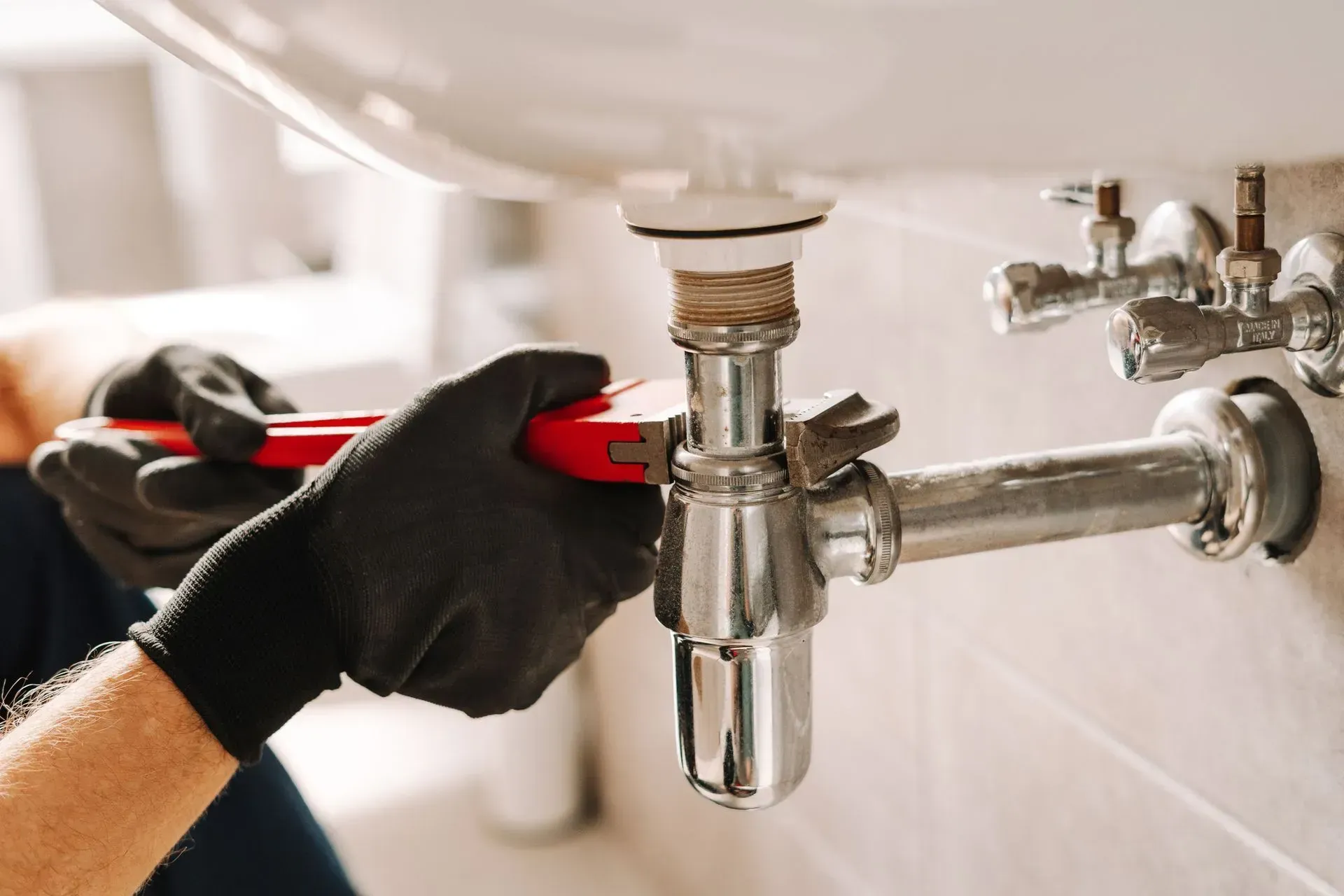 Gloved hands tightening a bathroom sink drain pipe under a white sink