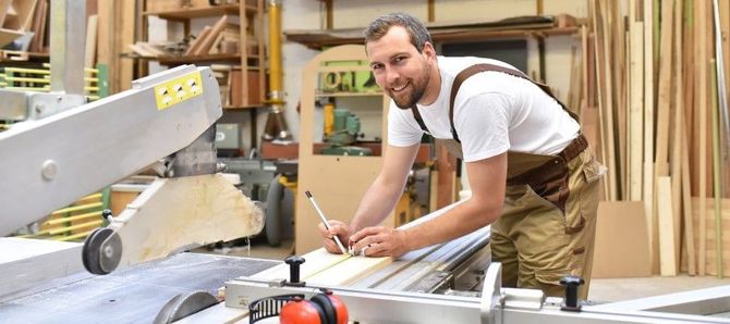Worker smiling at a table saw in a woodworking shop, with lumber stacked in the background