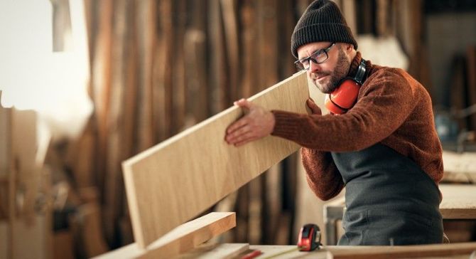 Woodworker in a beanie and safety glasses measures a light wood panel in a workshop.