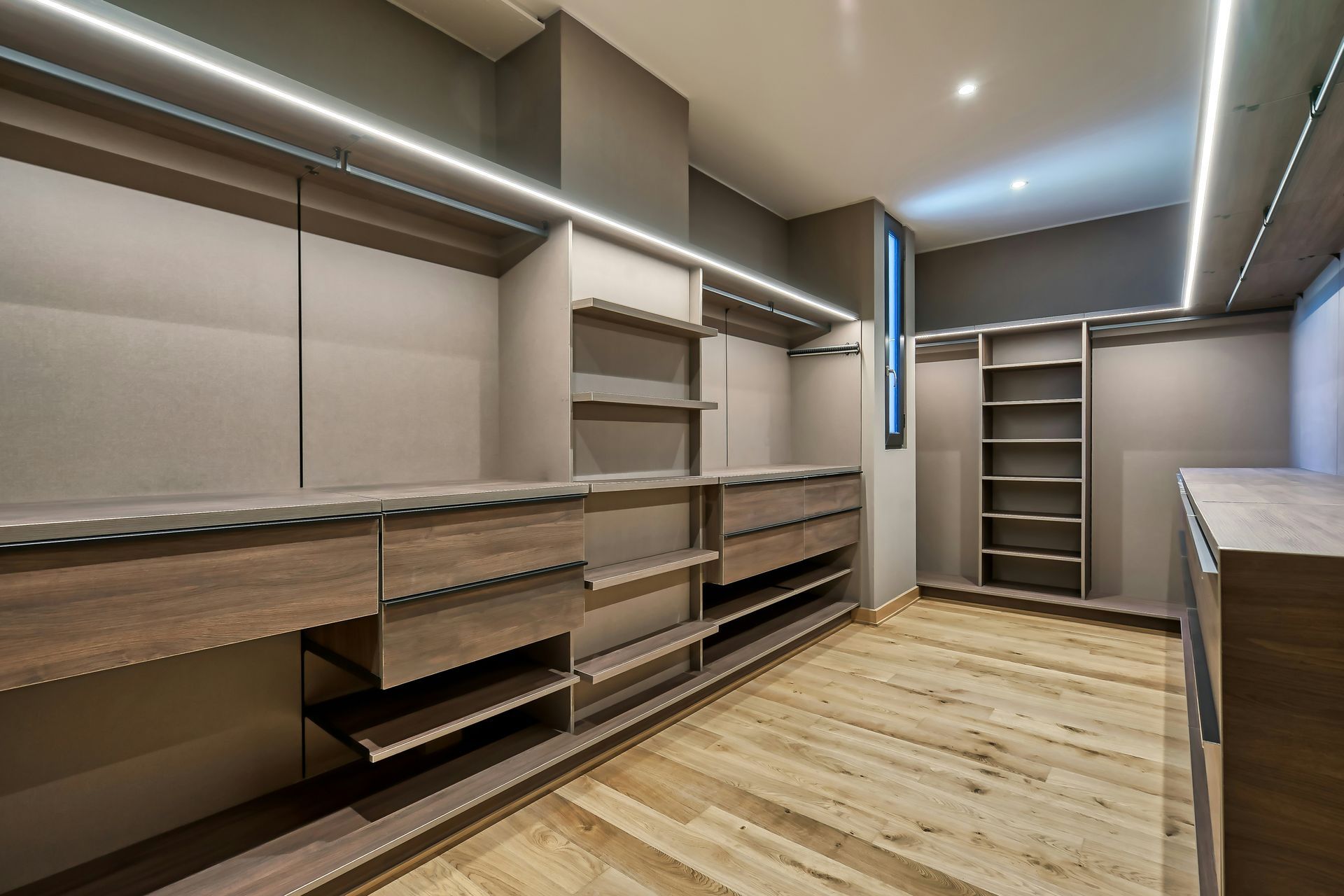 Empty walk-in closet with beige shelving, drawers, and light wood flooring under recessed lighting