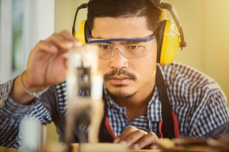 Man in safety glasses and yellow earmuffs examining a small object at a workbench