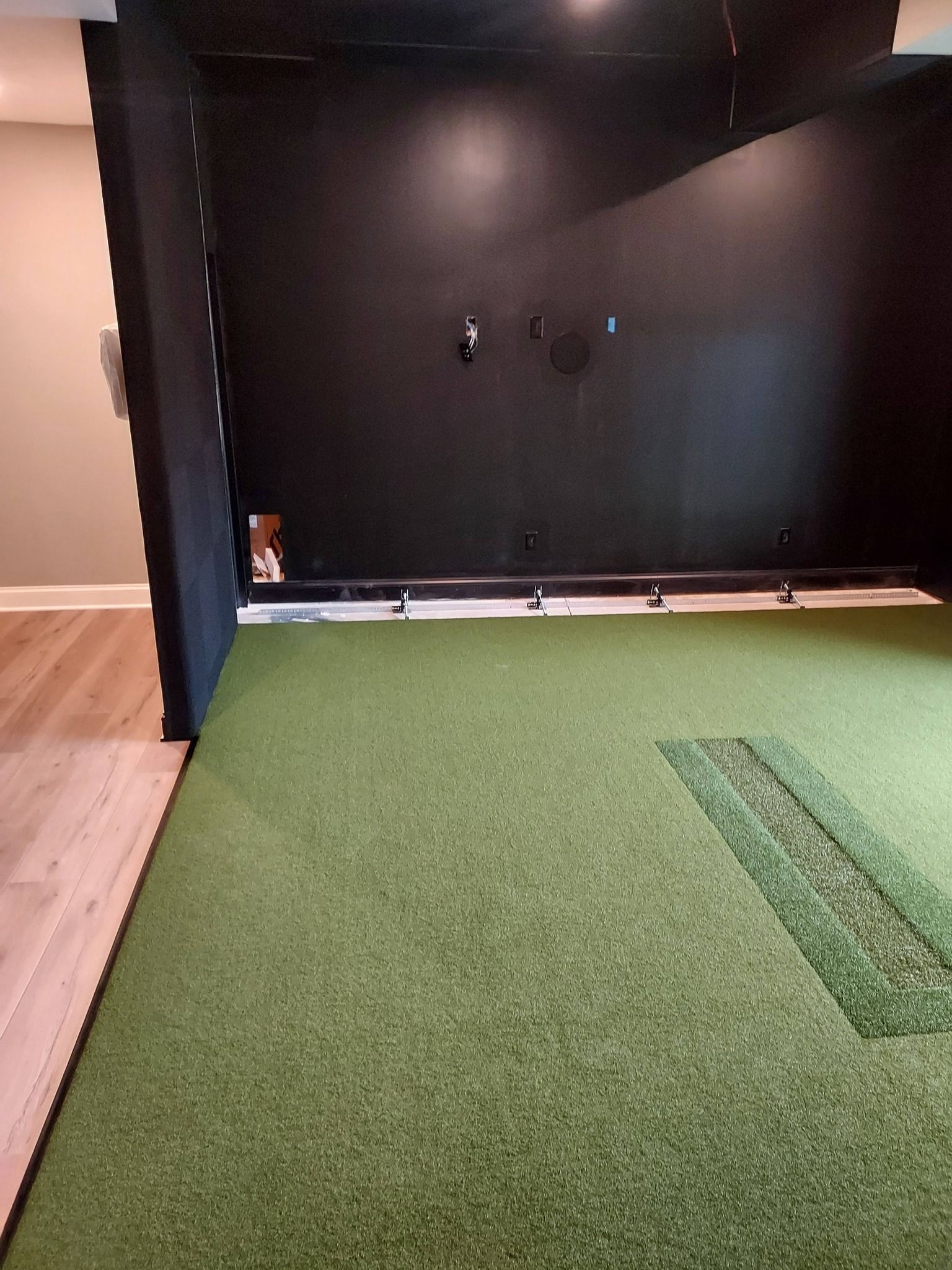 An indoor golf simulator room with a black impact screen, a green turf floor, and a hitting strip.