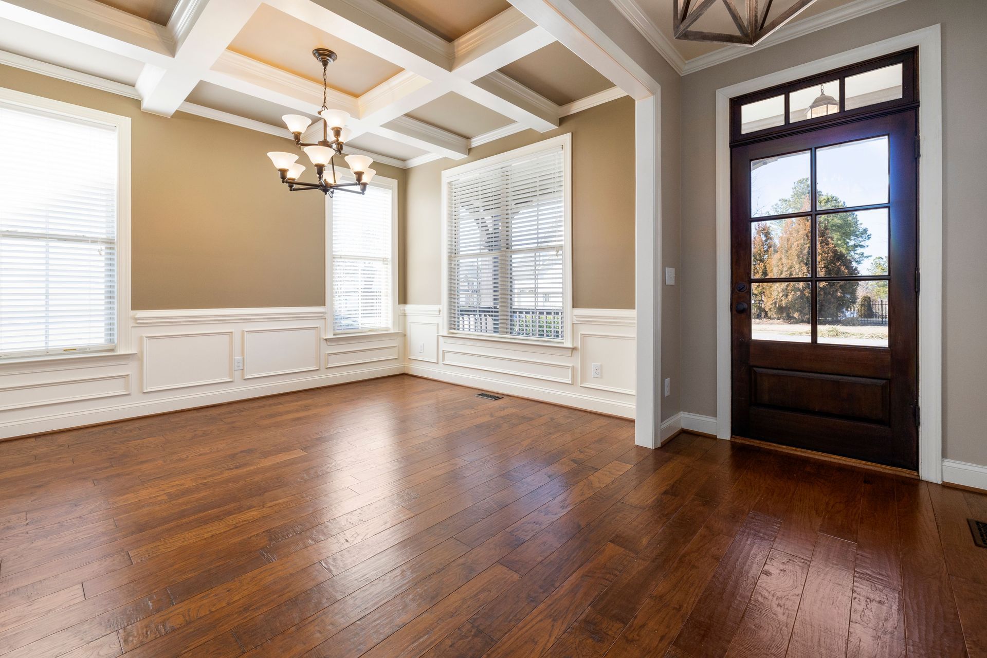 Empty dining room with hardwood floors, coffered ceiling, chandelier, white built-ins, and a dark front door.