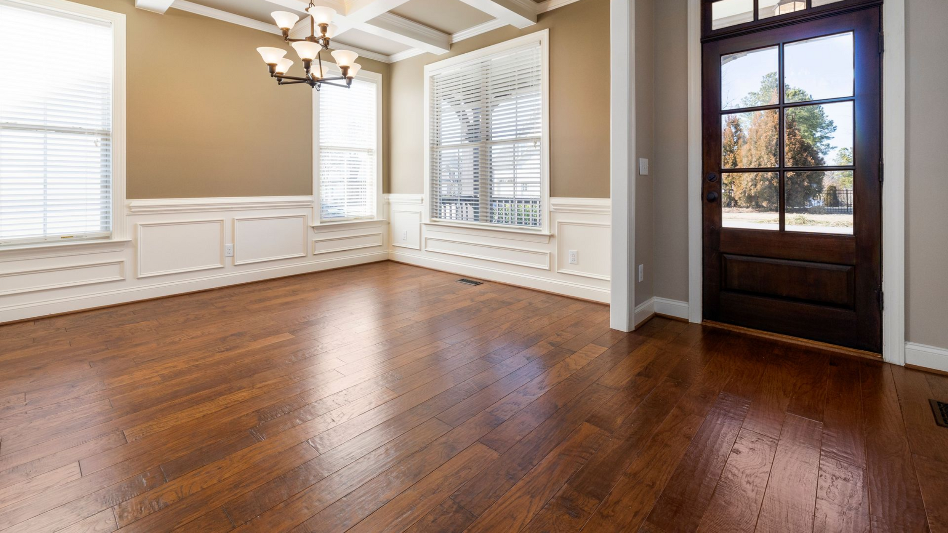 Empty dining room with hardwood floors, beige walls, white wainscoting, windows, and a dark wood front door