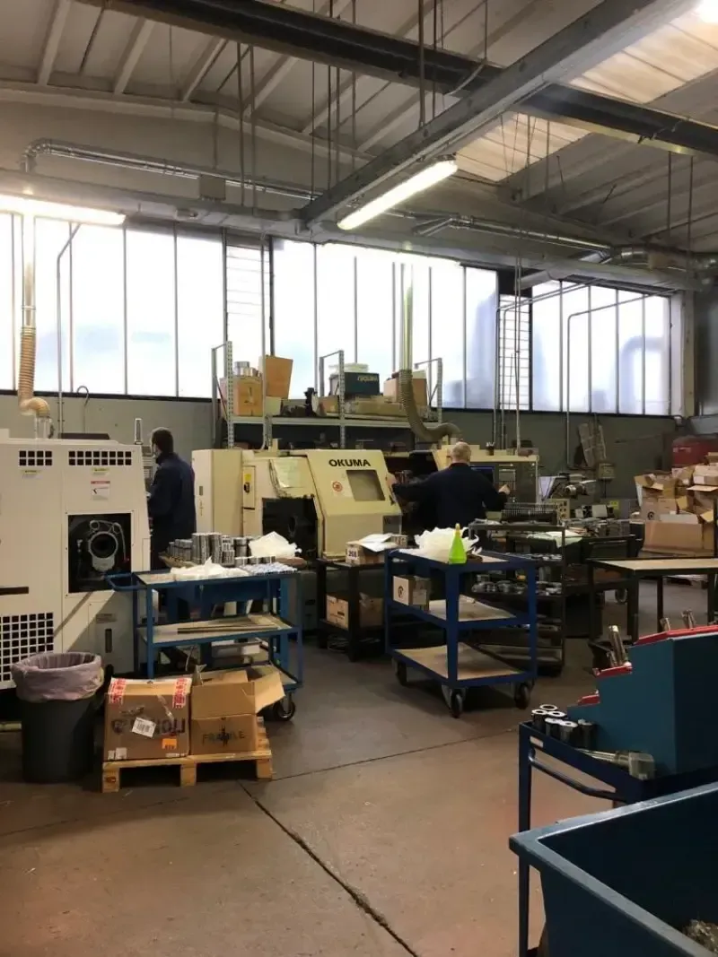 Stabilimento di produzione con macchinari e operai; interni luminosi, utensili, carrelli e finestre.