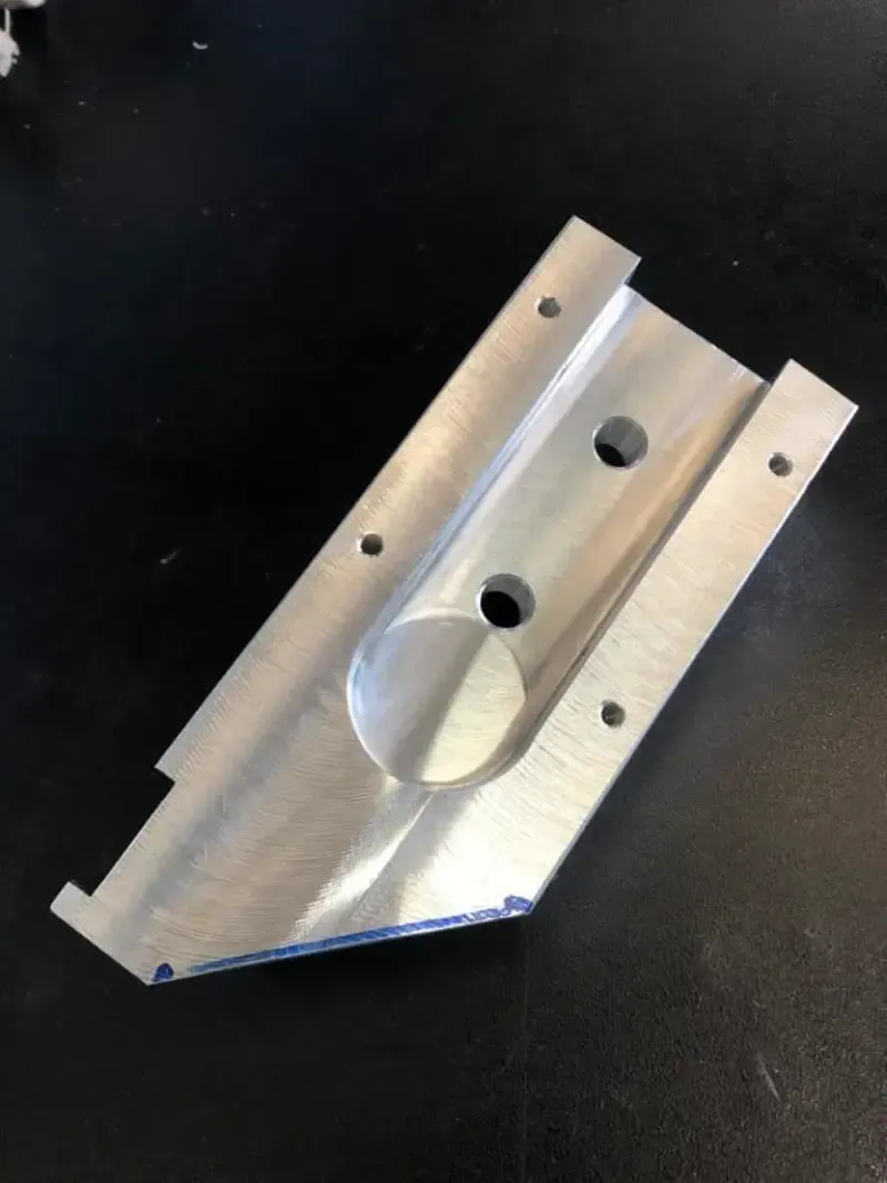 Pezzo metallico lavorato in alluminio con fori e fessure su una superficie nera.