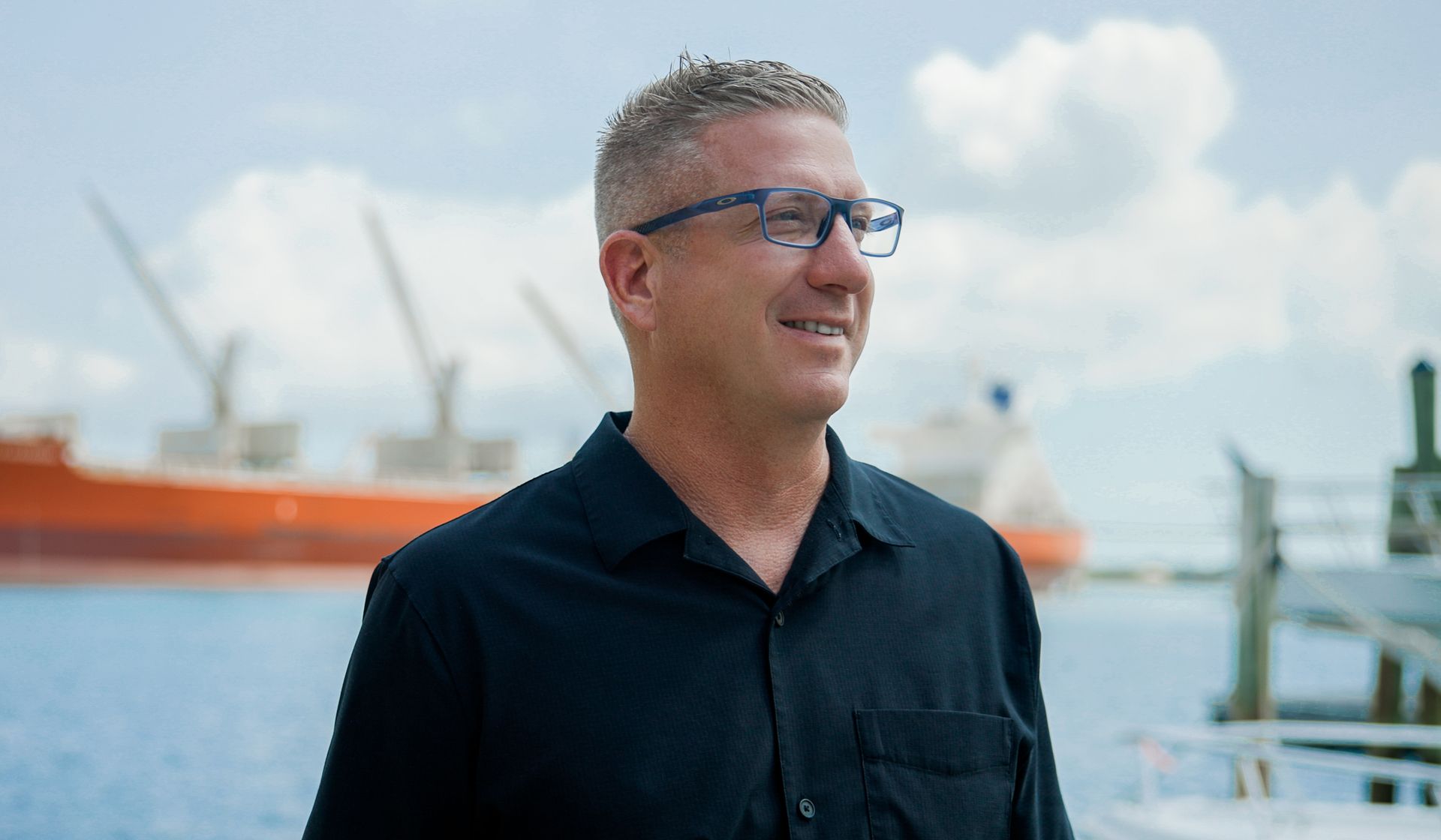 Man in glasses and dark shirt standing by the water with an orange ship in the background