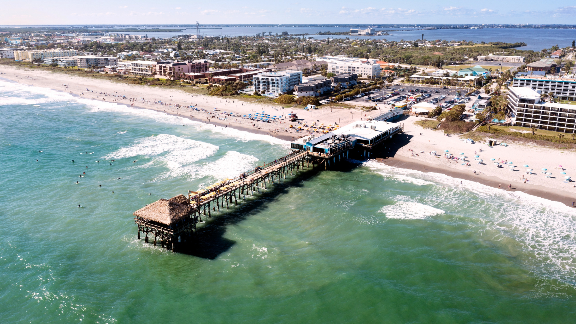 Aerial view of a long pier extending into turquoise water beside a sandy beach and coastal buildings.