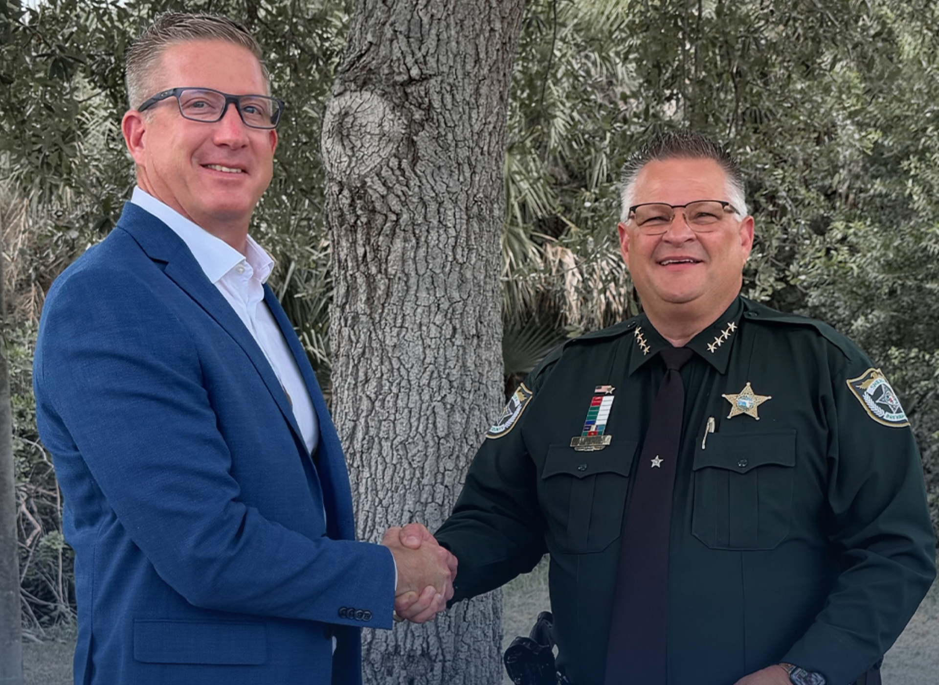 Two men shaking hands outdoors beside a tree, one in a blue suit and one in a sheriff’s uniform.