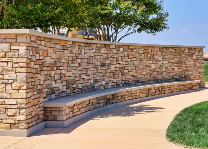 Curved stone wall with built-in bench beside a paved path and grass in a sunny outdoor park.
