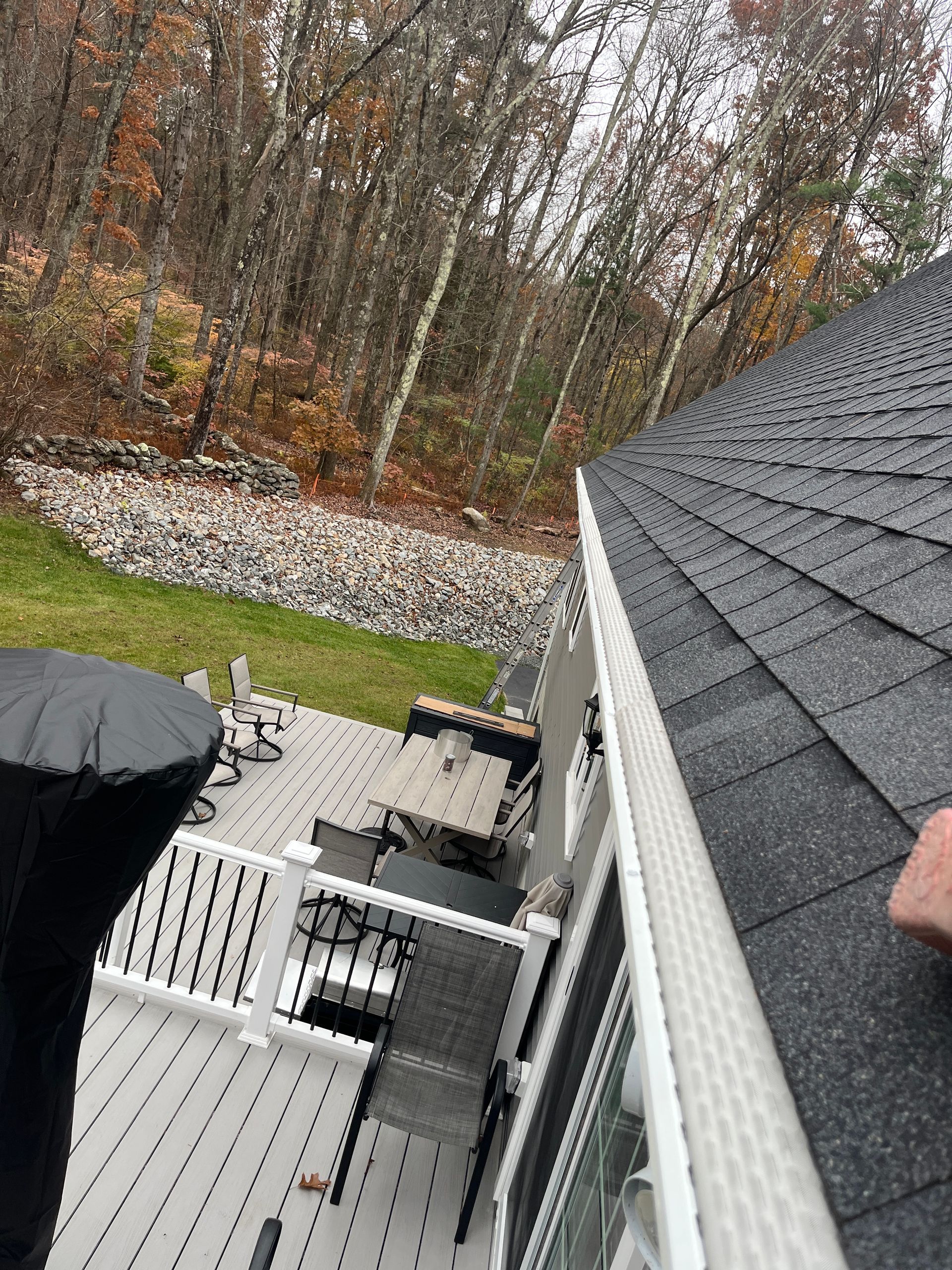 Backyard deck with white railing, black roof, patio furniture, and autumn trees beyond a rock border.