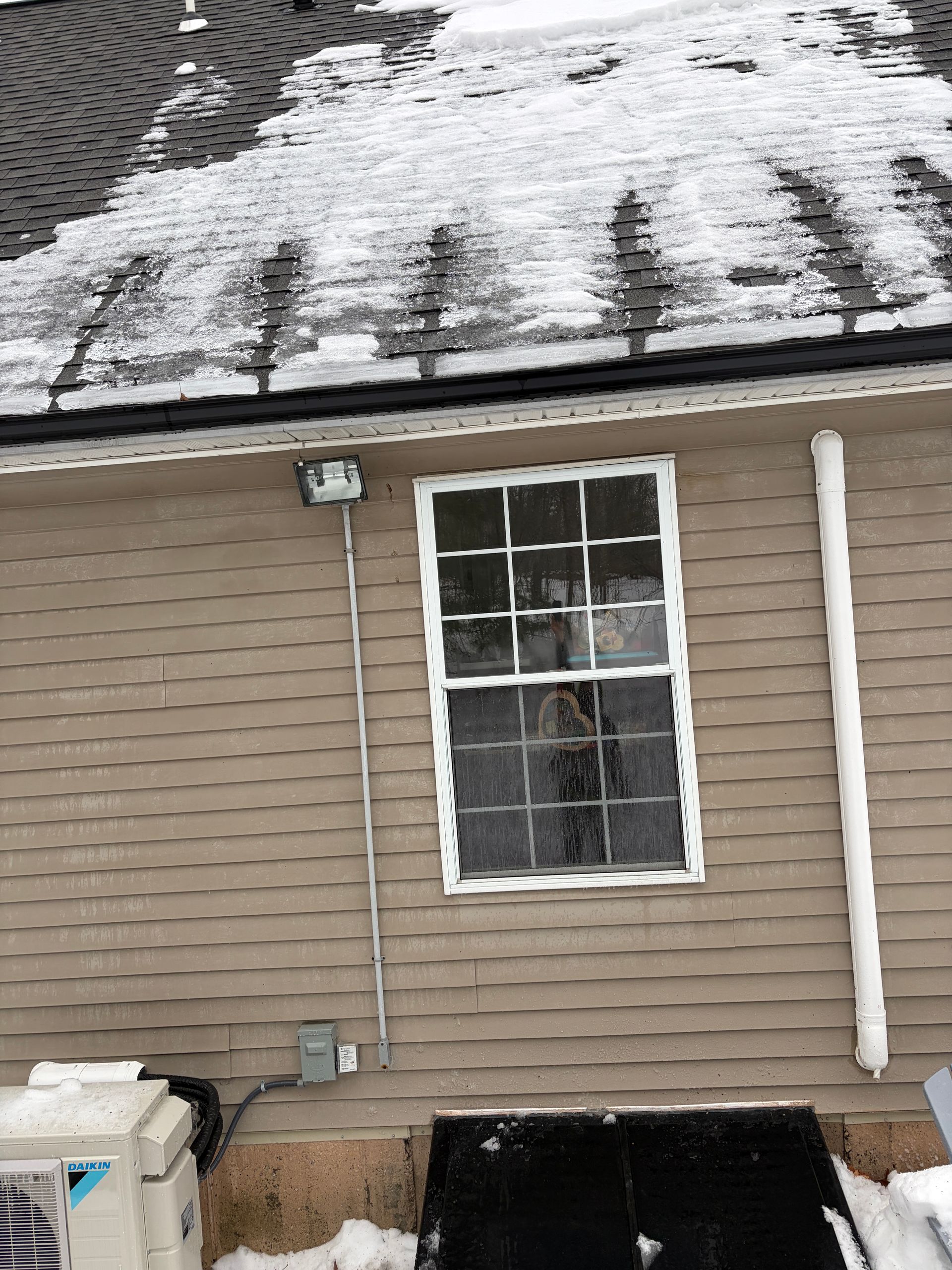 House exterior with tan siding, a white-framed window, and snow on the roof showing dark cleared patches.