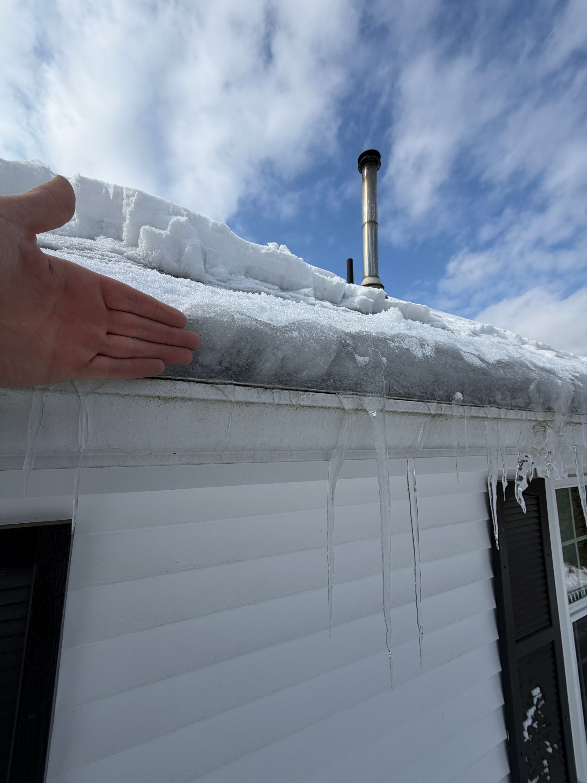 Hand next to thick snow piled on a roof above a white house with icicles and a blue sky