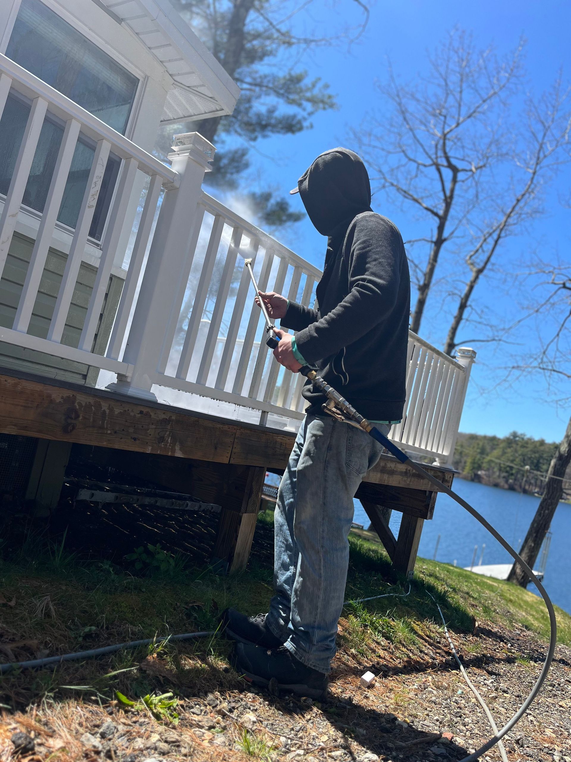 Person in black hoodie and jeans standing by a white porch, holding a tool near a lakeside house.