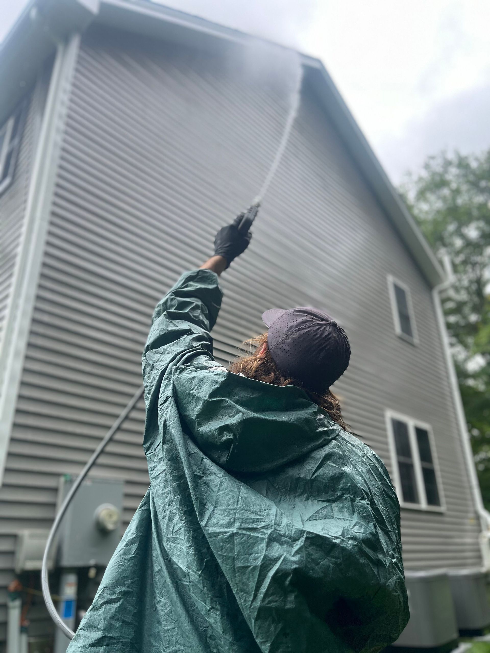 Person power-washing gray house siding from a ladder with a hose spray