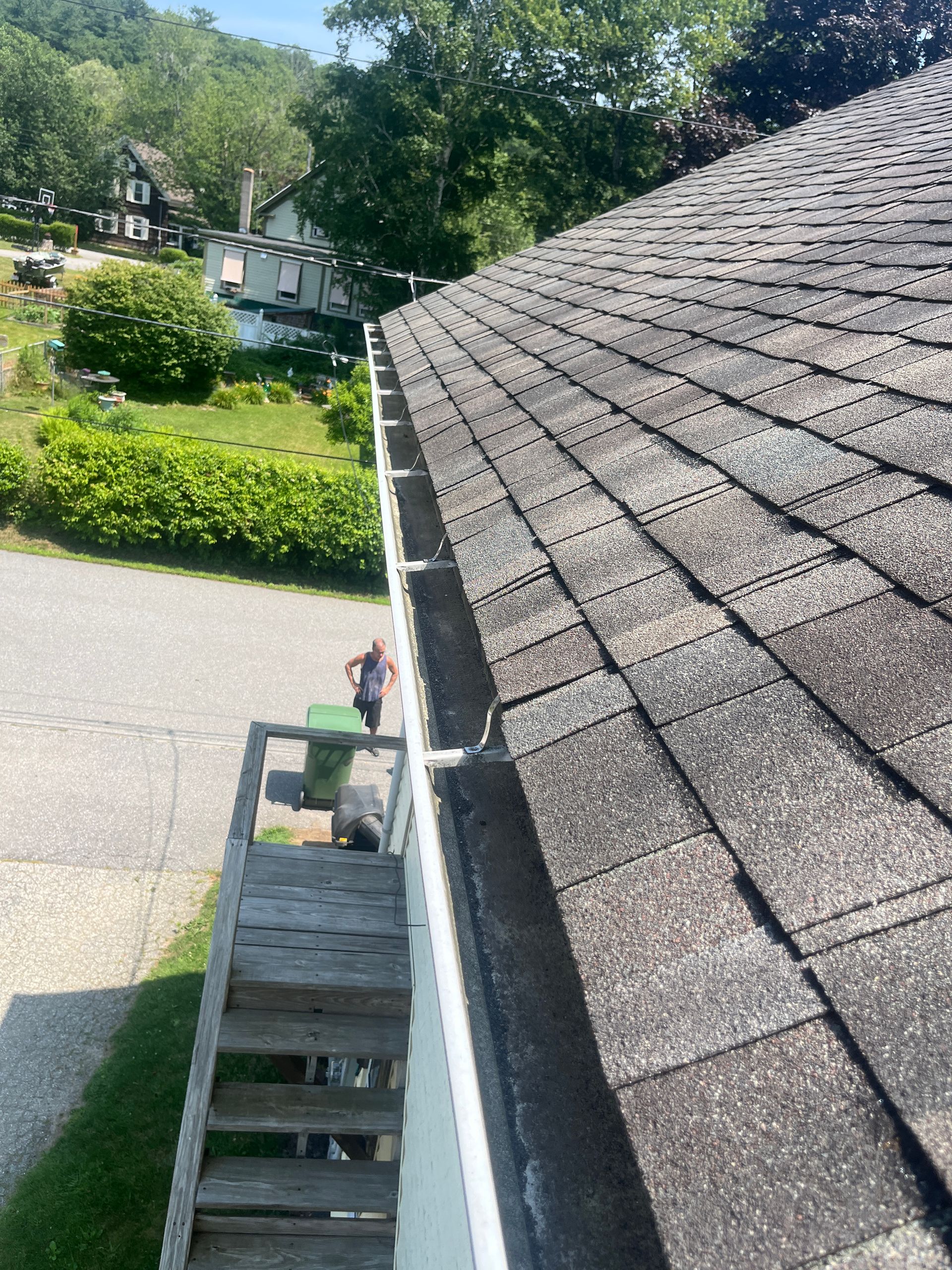 Roof with gray shingles and a gutter, viewed from a ladder beside a driveway and green yard.