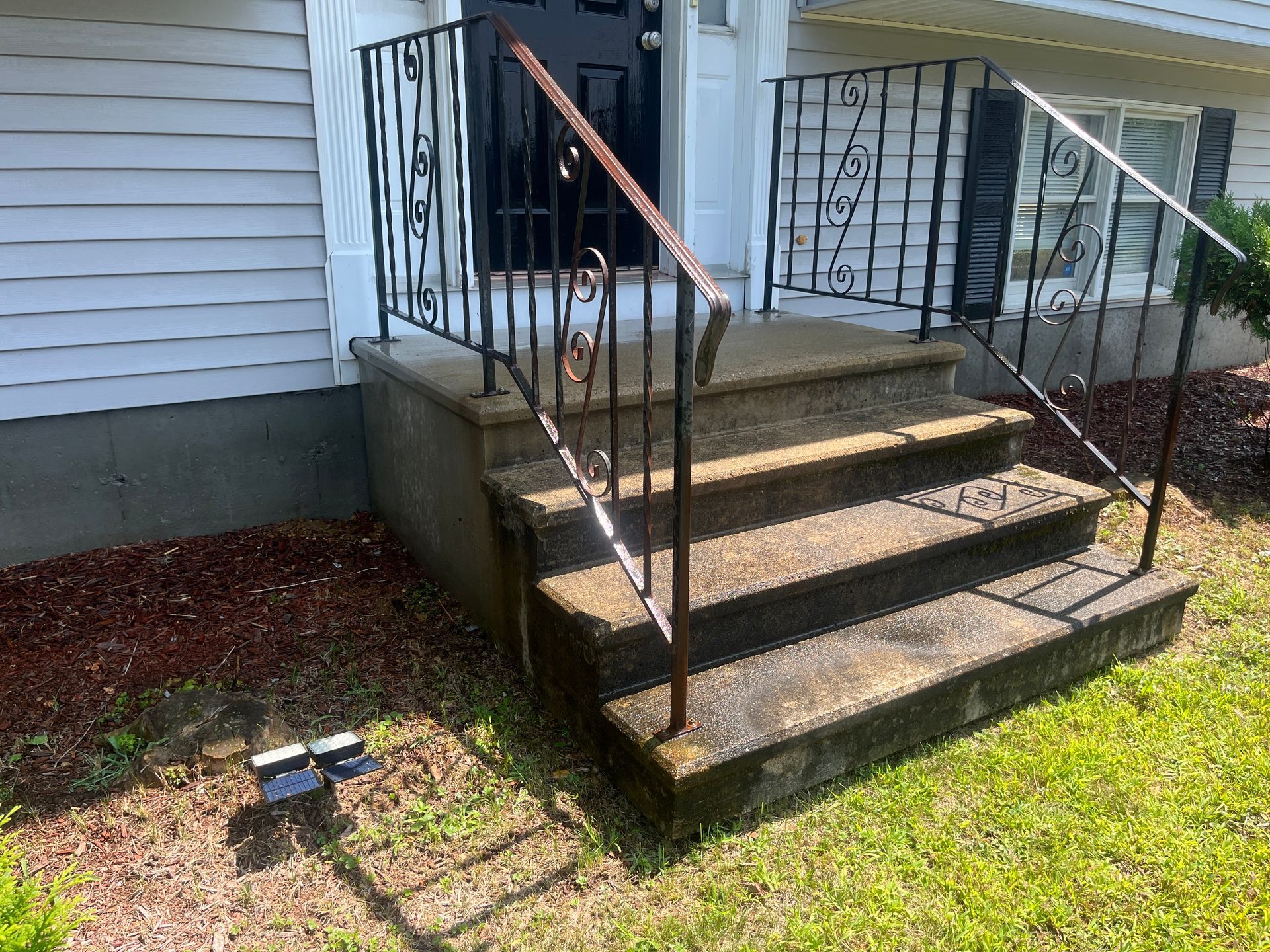Front steps of a house with black railings and a concrete landing, beside a small lawn light.