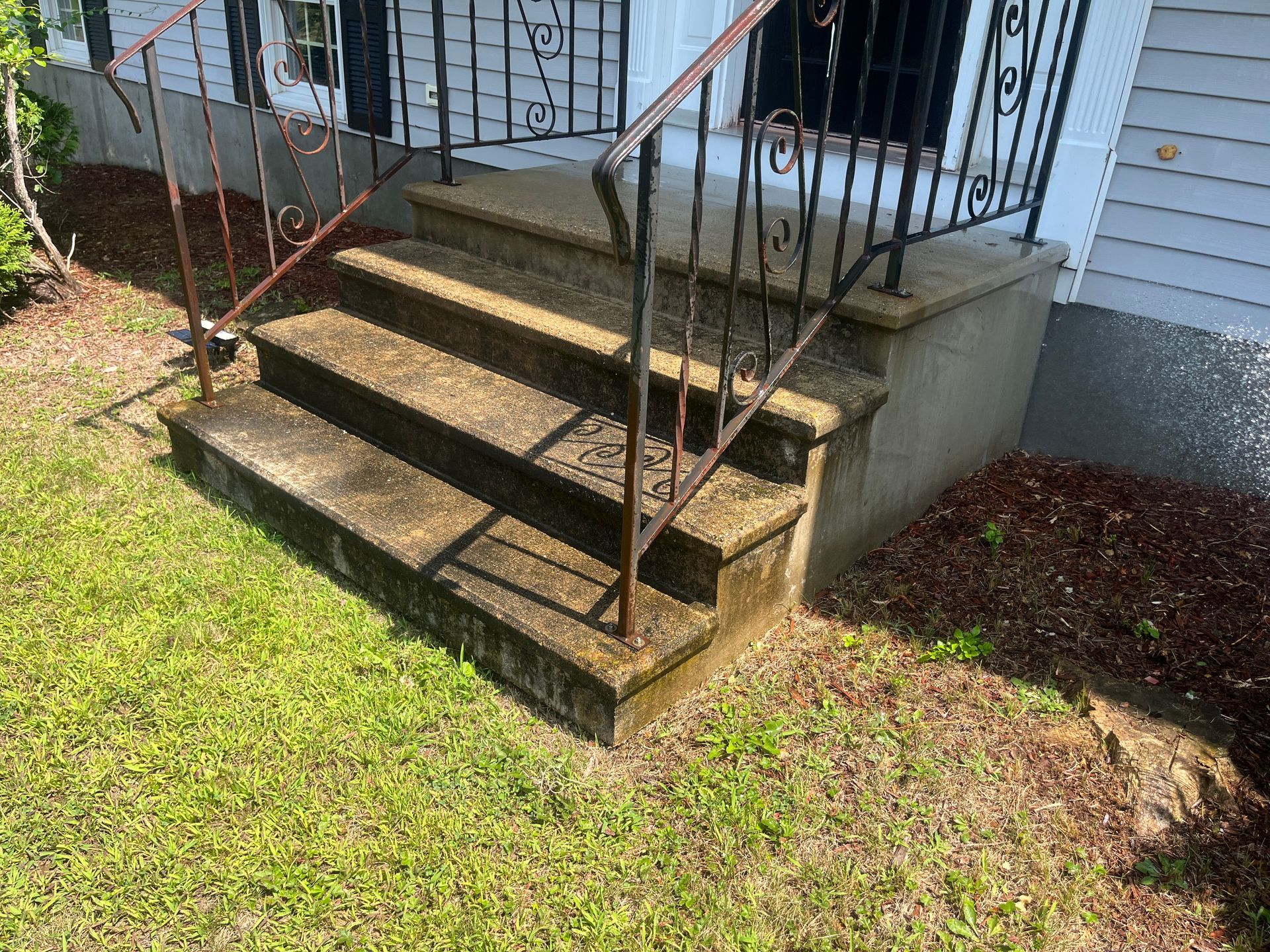 Front steps with metal railings leading to a porch beside a white house and grassy yard.